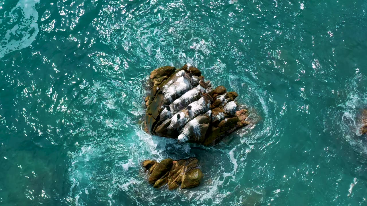 Overhead view at Secrets Marquis Los Cabos, Mexico, showing the main rock high in frame with white foam and a smaller rock below it in clear blue-green water, with no visible people.