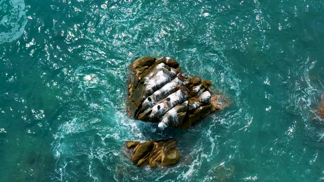 Overhead frame from Secrets Marquis Los Cabos, Mexico, with teal water pushing foam against the left and upper sides of the rock while the smaller rock sits below, with no visible people.