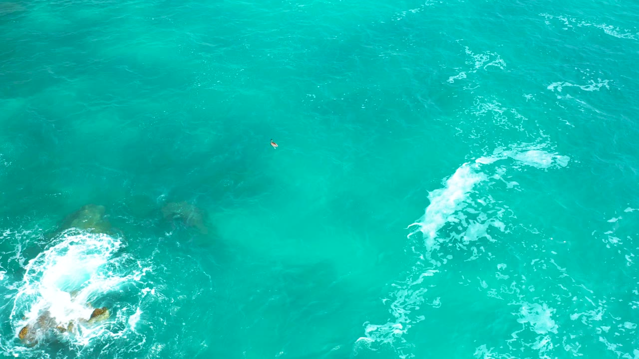 Overhead view at Secrets Marquis Los Cabos, Mexico, where a small rocky patch sits near the lower-left edge and white foam trails through bright turquoise water, with no visible people.