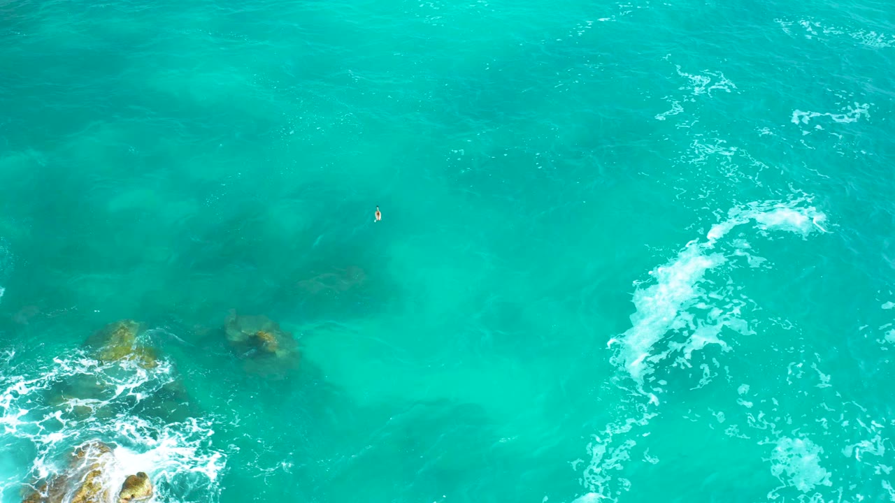 Overhead Los Cabos ocean frame with a curved white foam line crossing the turquoise water and rock edges visible at the bottom, no visible people.