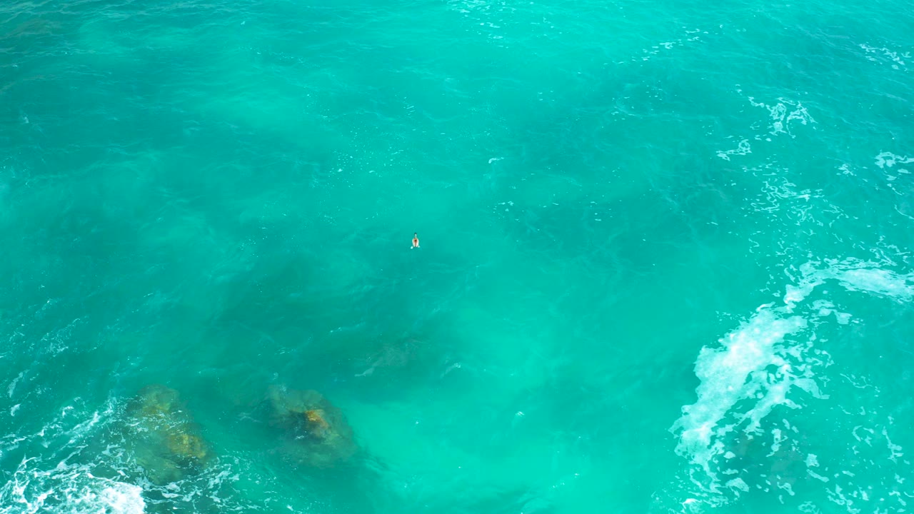 Overhead view of green turquoise seawater at Secrets Marquis Los Cabos, Mexico, with submerged rocks low in frame, white foam crossing the center, and no visible people.