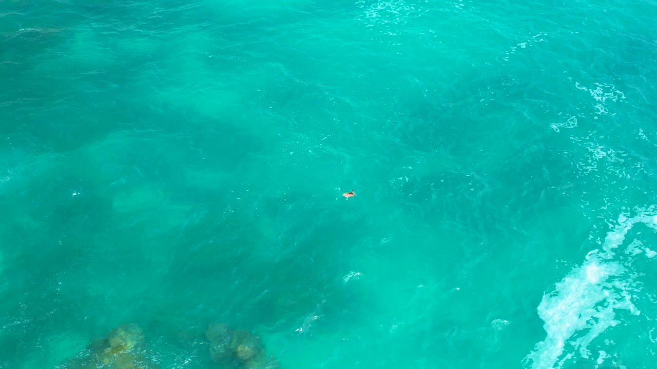 Top-down turquoise ocean with submerged rocks and white foam at the right edge.