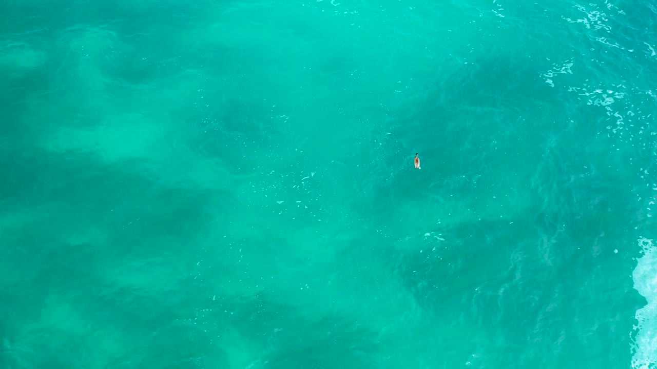 Wide overhead view of the green-blue sea at Secrets Marquis Los Cabos, Mexico, with one tiny swimmer near the upper right and pale foam just inside the right edge.