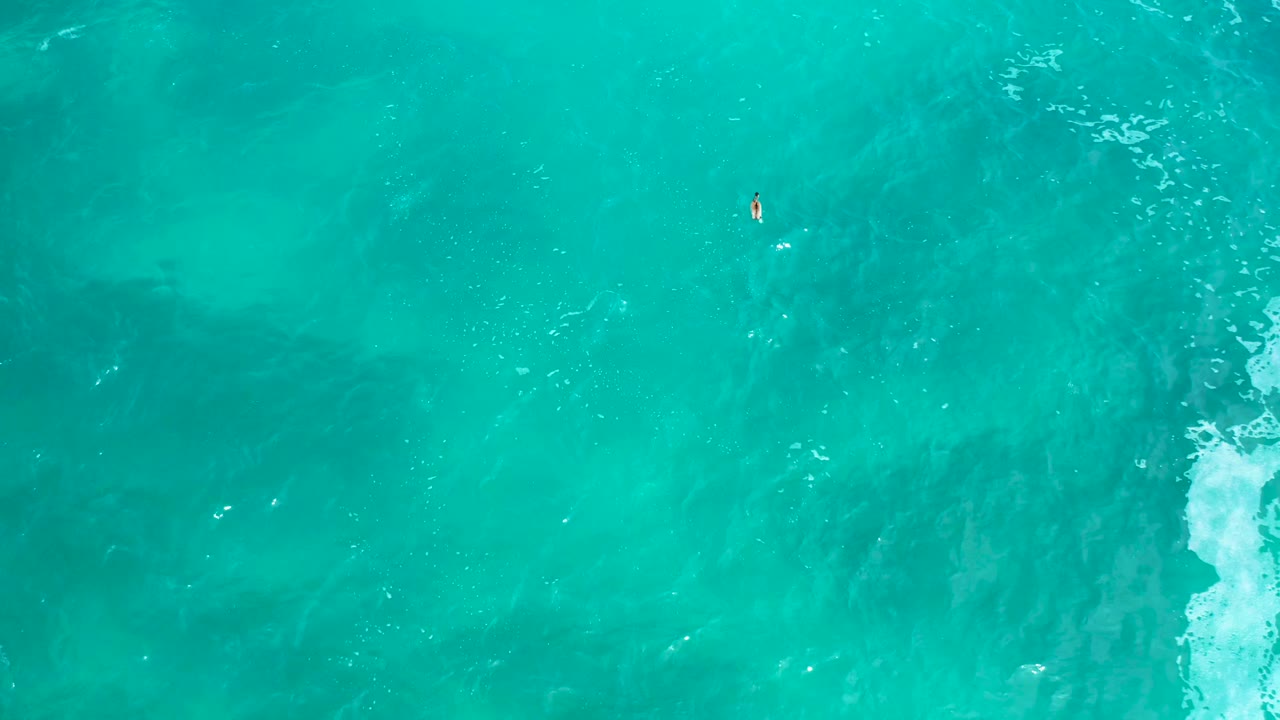 Aerial view over turquoise water at Secrets Marquis Los Cabos, showing a tiny drifting surface speck above center, faint ripples, darker sea tones, and no visible people.