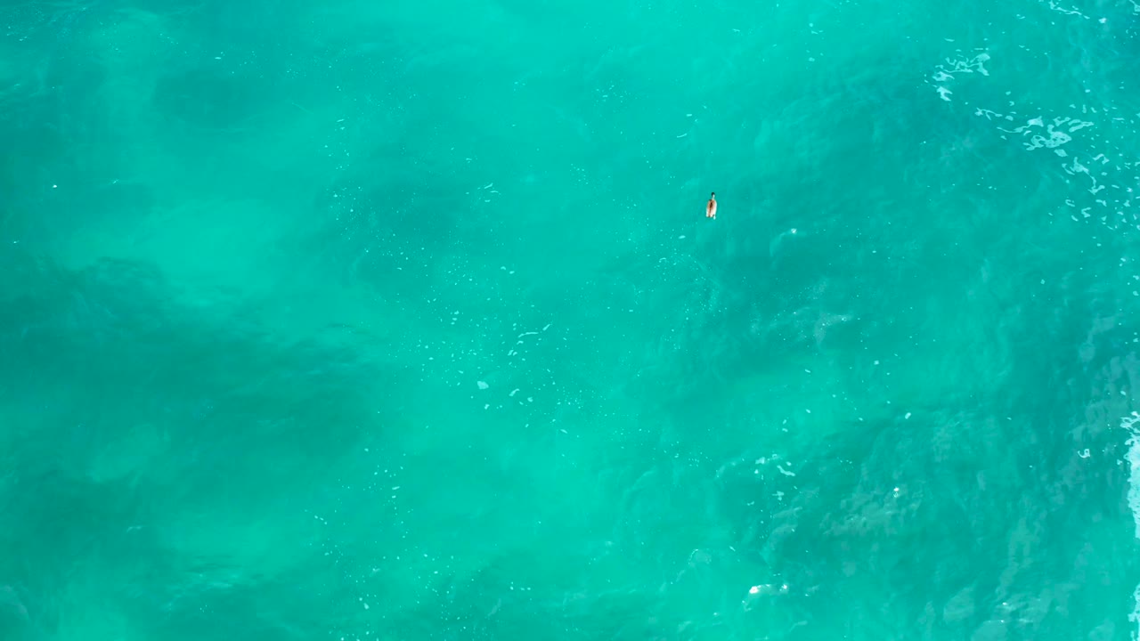 Overhead blue-green ocean at Secrets Marquis Los Cabos, with soft darker shapes under the surface, a tiny floating speck near the upper center, and no visible people.