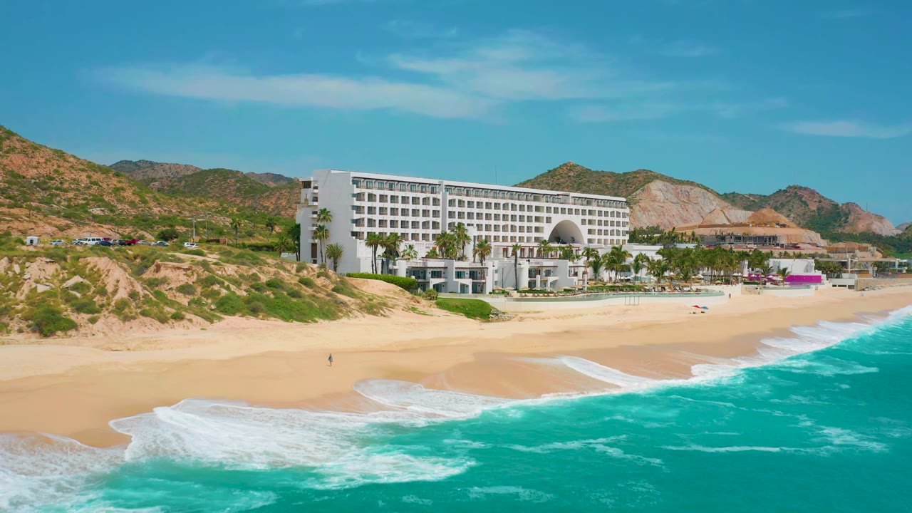 Elevated resort beach view at Secrets Marquis Los Cabos in Mexico, with foamy surf spreading farther across the lower sand and the white hotel centered behind palms, no visible people.