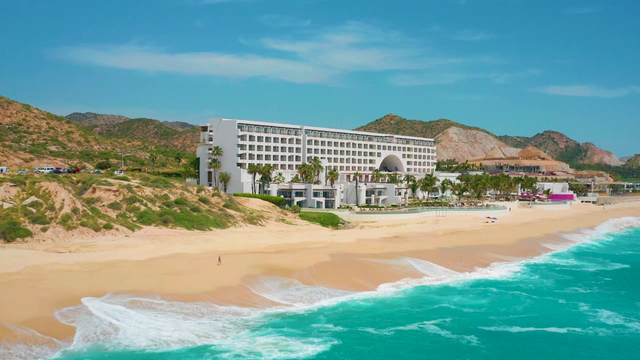 Wide aerial-style frame of the Secrets Marquis Los Cabos resort in Mexico, with the hotel slightly right of center, palm trees along the beach, and bright turquoise water filling the lower right, no visible people.
