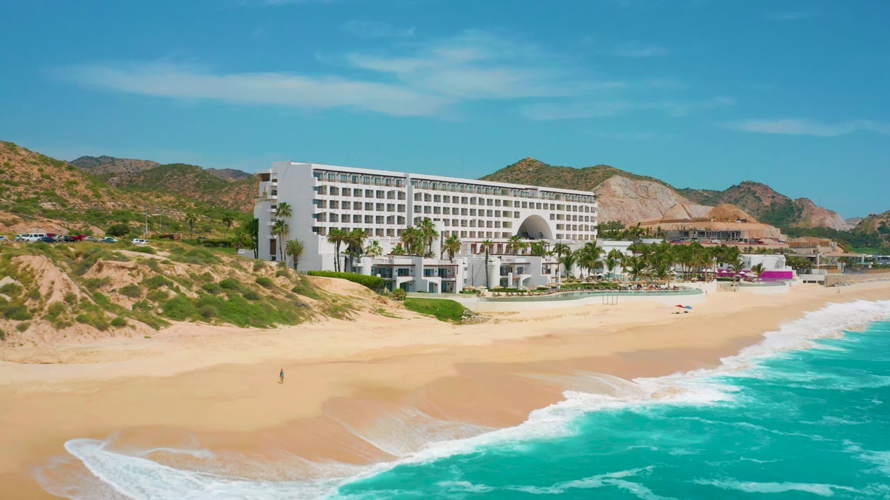 Secrets Marquis Los Cabos in Mexico seen from an elevated beach angle, with a broad open sand foreground, the white resort and palms in the midground, and waves entering from the right, no visible people.