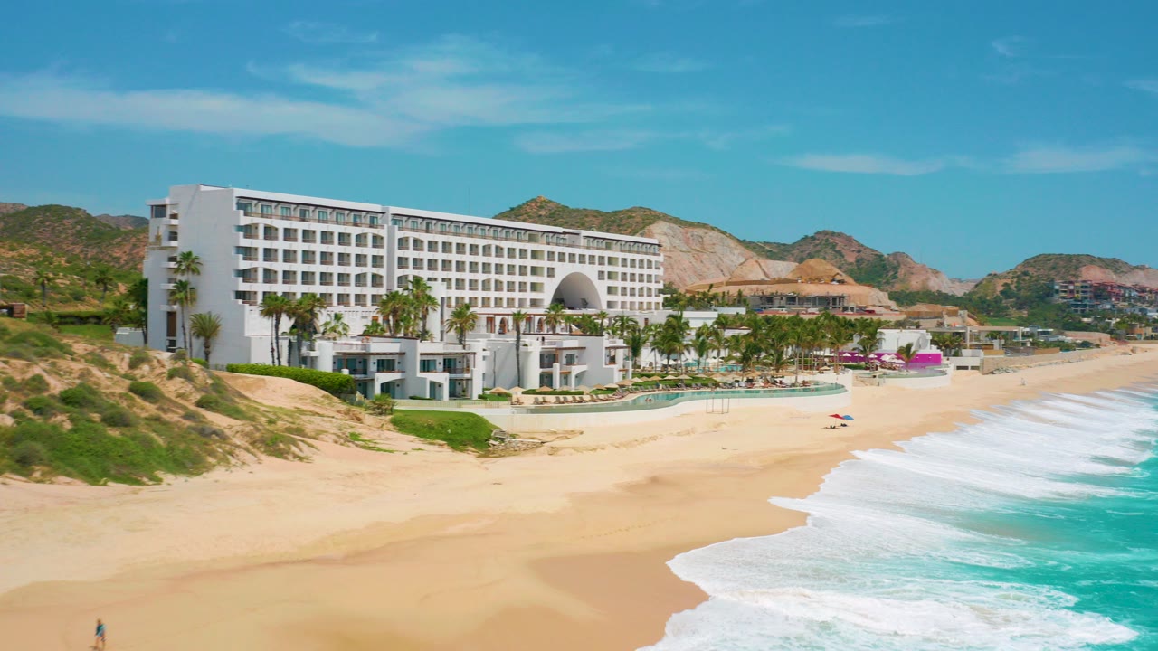 At 0:00, a wide aerial view shows the white Secrets Marquis Los Cabos resort above a sandy beach, turquoise surf on the right, low hills in back, and no visible people.