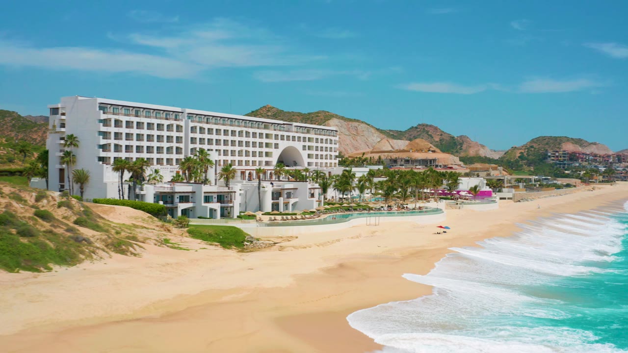 At 0:02, the elevated view frames the resort facade, palm-lined grounds, pale sand, and bright blue water with a smooth foam band along the shore and no visible people.