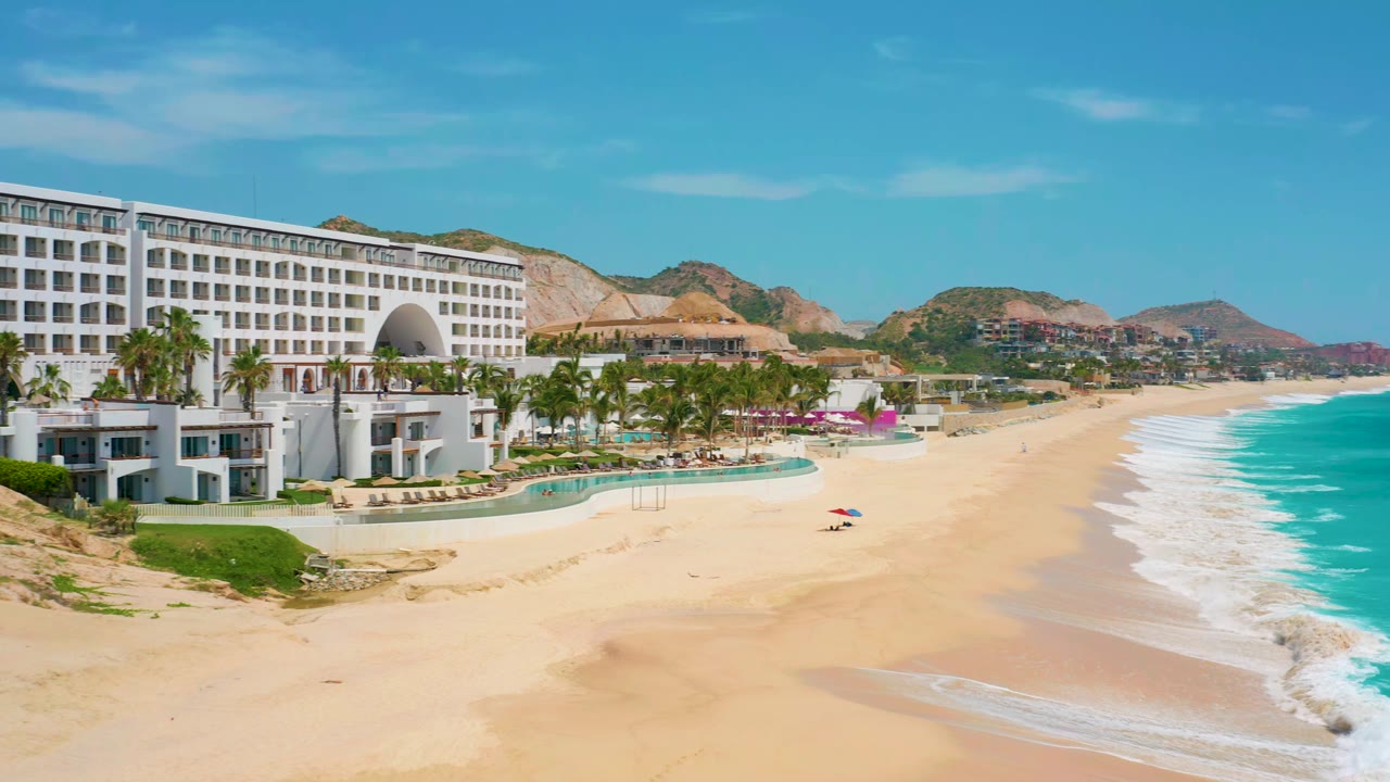 White beachfront resort, palm trees, sand, waves, and hills under blue sky.