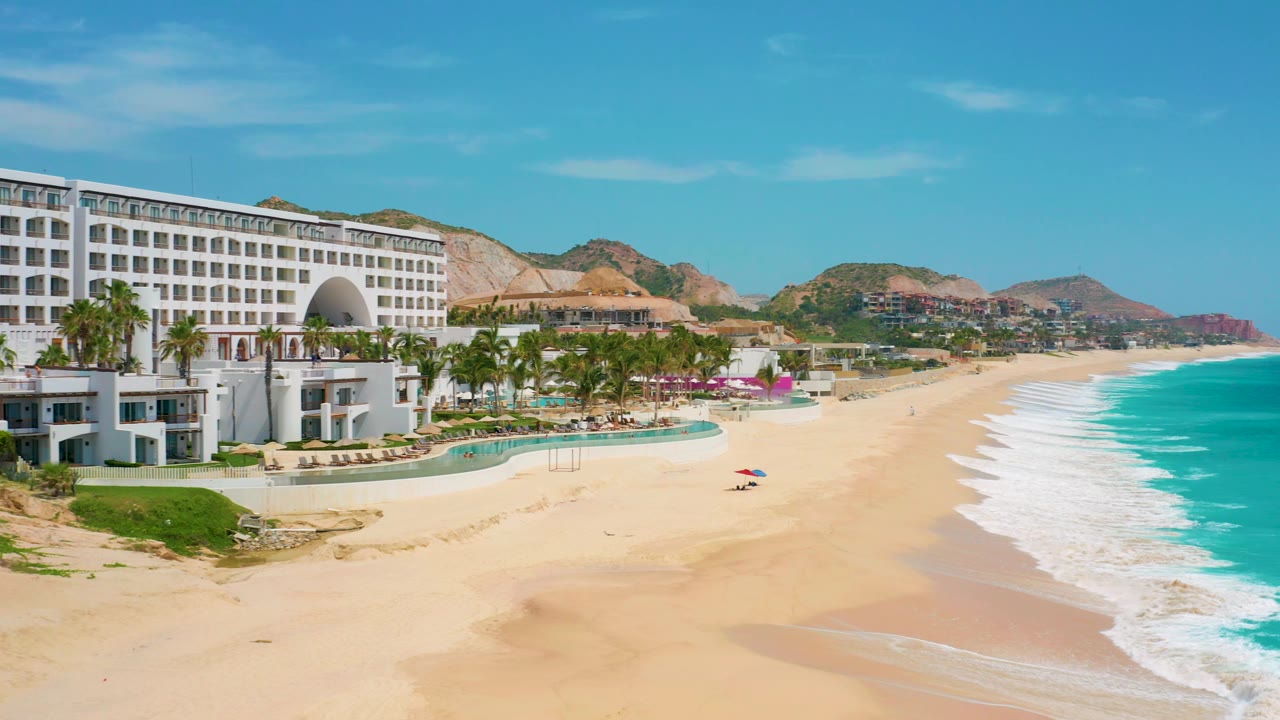 Wide frame of Secrets Marquis Los Cabos in Mexico showing the resort at left, a curving pale beach, turquoise water, white wave bands, distant hills, and no visible people.
