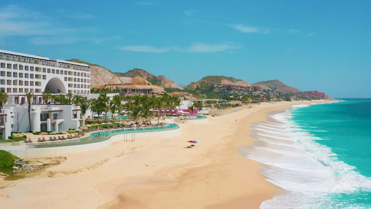 Wide aerial frame of Secrets Marquis Los Cabos in Mexico with a white resort building at left, palms and pool terraces below, a broad sandy beach, turquoise surf, distant hills, and tiny people visible on the sand.