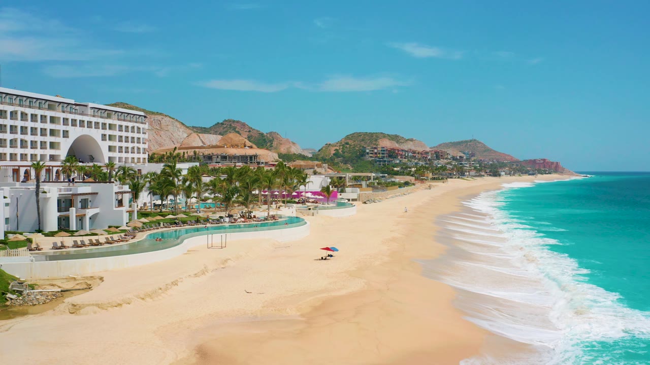 Aerial frame of the Los Cabos resort coastline with the building cropped along the left side, pools and palms in the foreground, a small red and blue beach umbrella on the sand, turquoise water to the right, hills behind, and tiny people visible.