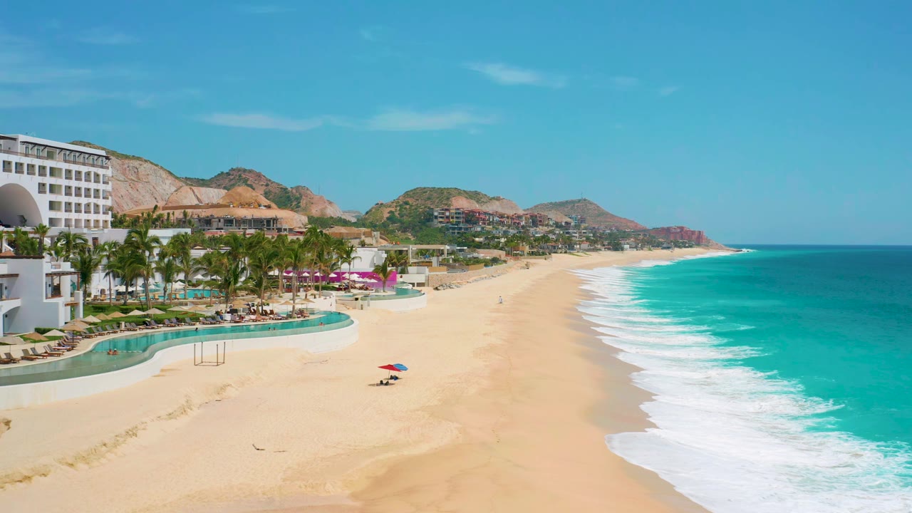 One second into the clip, the Secrets Marquis Los Cabos beachfront remains wide in frame with pools and palms at left, foamy waves at right, hills beyond, and no visible people.