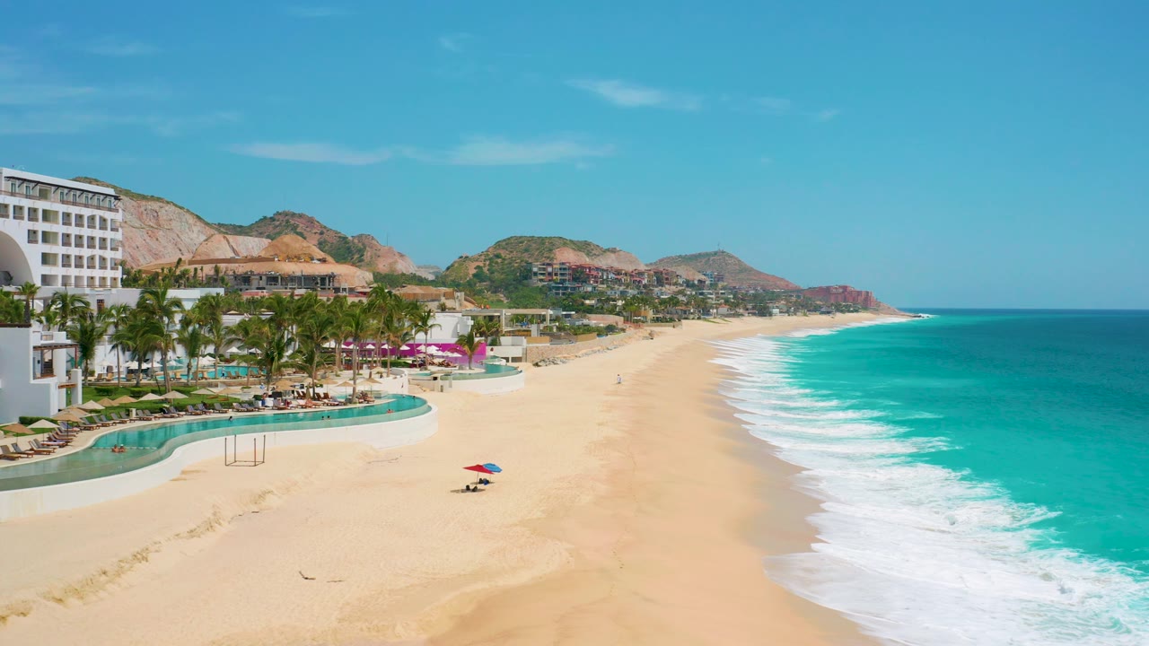 At 0:02, the Mexico resort frontage sits at the left edge while the bright sand, turquoise water, white foam bands, distant hills, and no visible people fill the rest of the view.