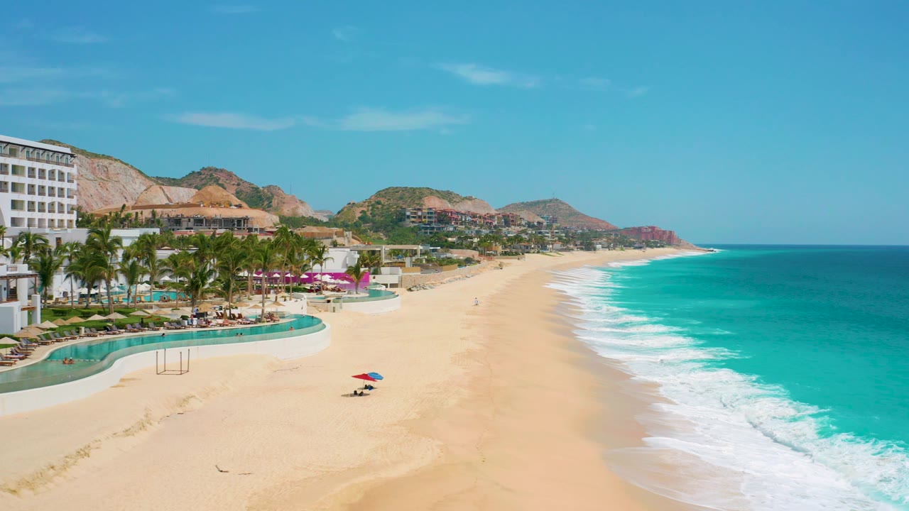 At 0:03, the elevated view shows the resort pool curves and palms more prominently at left, with open sand, breaking surf, hills in back, and no visible people.