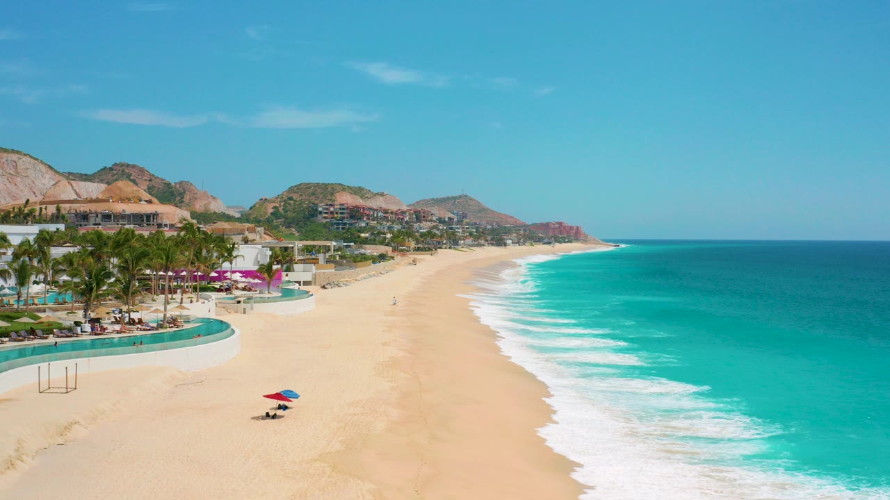 Aerial frame of Secrets Marquis Los Cabos, Mexico showing turquoise waves beside a broad sandy beach, resort pools and palms at left, distant hills, and tiny beachgoers near a red and blue umbrella.