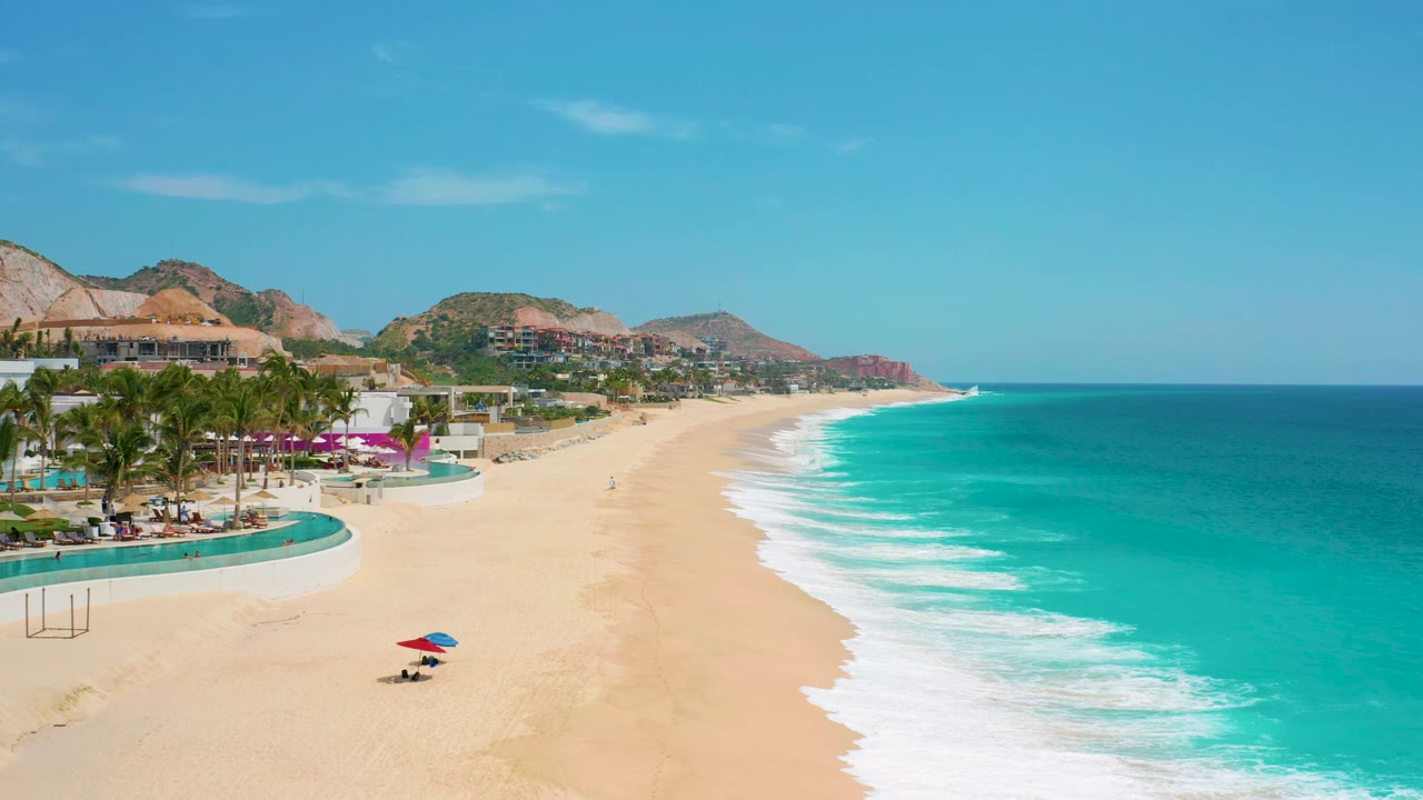 A bright elevated view of Secrets Marquis Los Cabos, Mexico shows open tan sand, bands of white foam, deep turquoise ocean, resort landscaping at left, and small beachgoers near the lower beach.