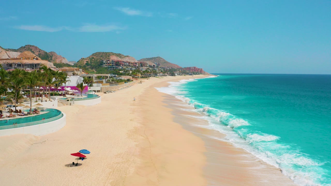 Aerial view at Secrets Marquis Los Cabos, Mexico, with resort pools and palms on the left, a broad sandy beach below, turquoise ocean on the right, and small people visible near umbrellas and along the shore.