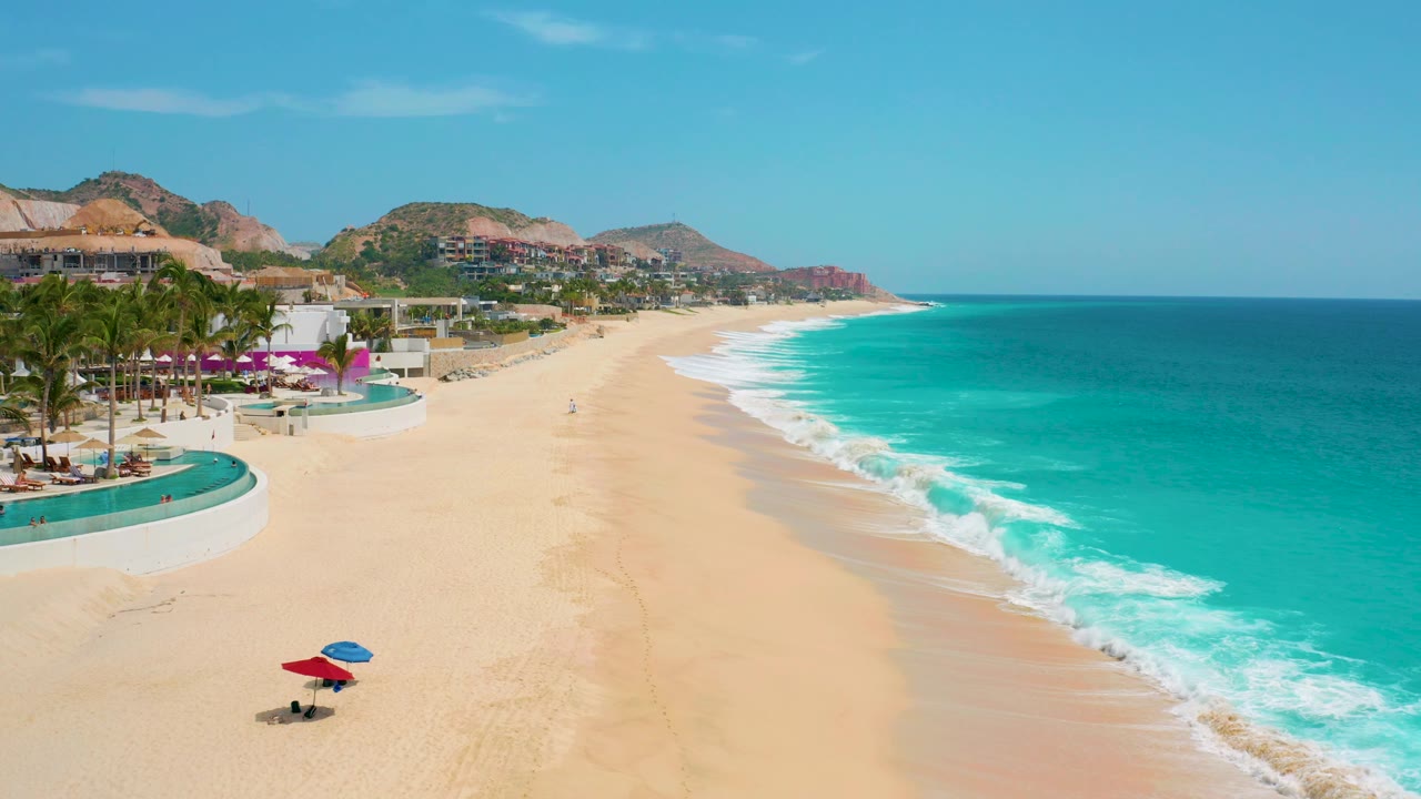 Aerial view over Secrets Marquis Los Cabos, Mexico, with the resort edge and palm-lined pool area at left, open sand in the center, bright surf on the right, and tiny people visible on the beach.