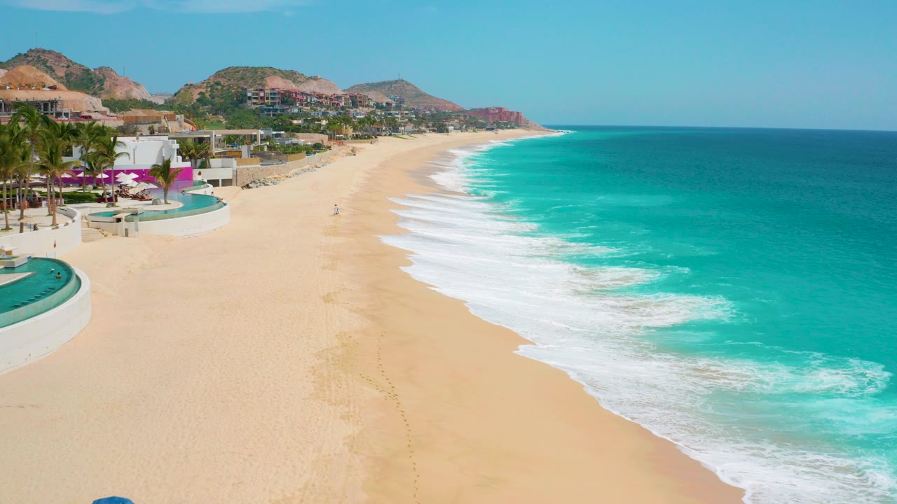 Opening aerial frame of Secrets Marquis Los Cabos beach in Mexico, with pale sand below, turquoise surf to the right, resort buildings and palms to the left, and no recognizable people visible.