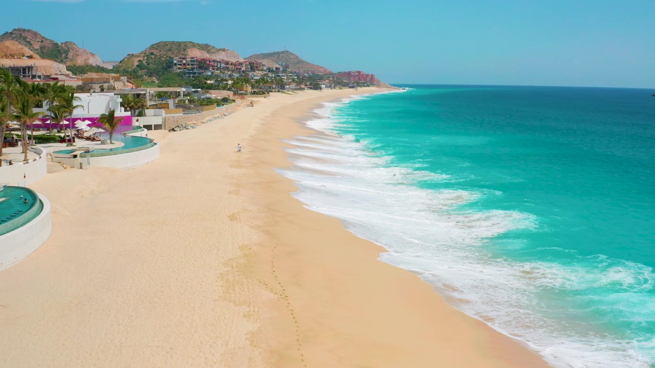 Aerial frame at Secrets Marquis Los Cabos in Mexico with a curved pool edge and palms at left, broad sand through the center, turquoise ocean at right, and no recognizable people visible.