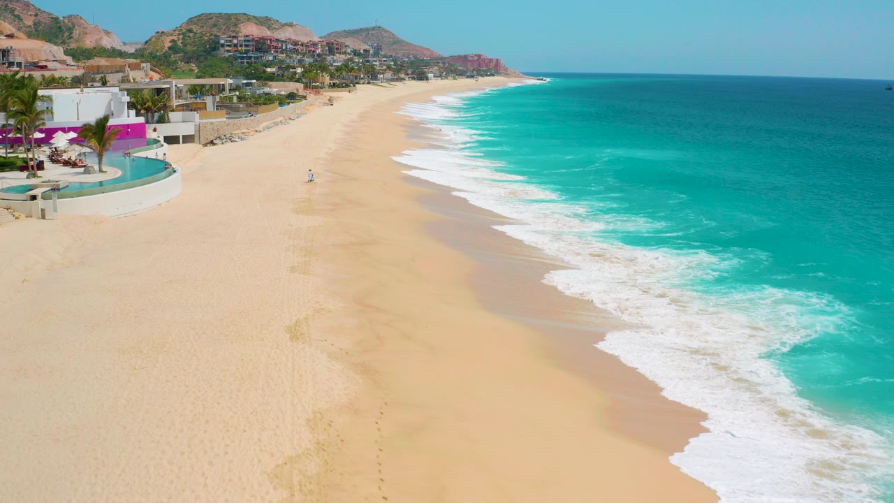 Wide aerial frame of Secrets Marquis Los Cabos in Mexico, with pale sand, turquoise water, white surf, resort buildings at left, coastal hills beyond, and no visible people.