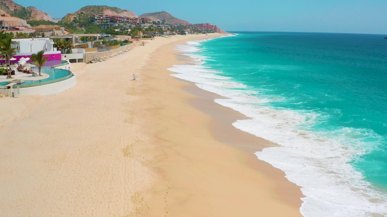 Aerial frame from Secrets Marquis Los Cabos showing the beach curving away, bright blue-green ocean to the right, white foam bands at shore, buildings and hills to the left, and no visible people.