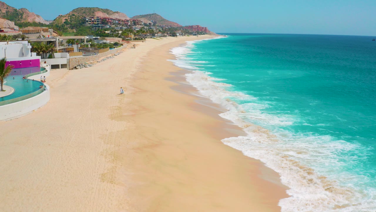 Opening view over the Secrets Marquis Los Cabos beach in Mexico, with turquoise water at right, pale sand below, resort buildings at left, low hills in back, and a tiny distant beachgoer on the sand.