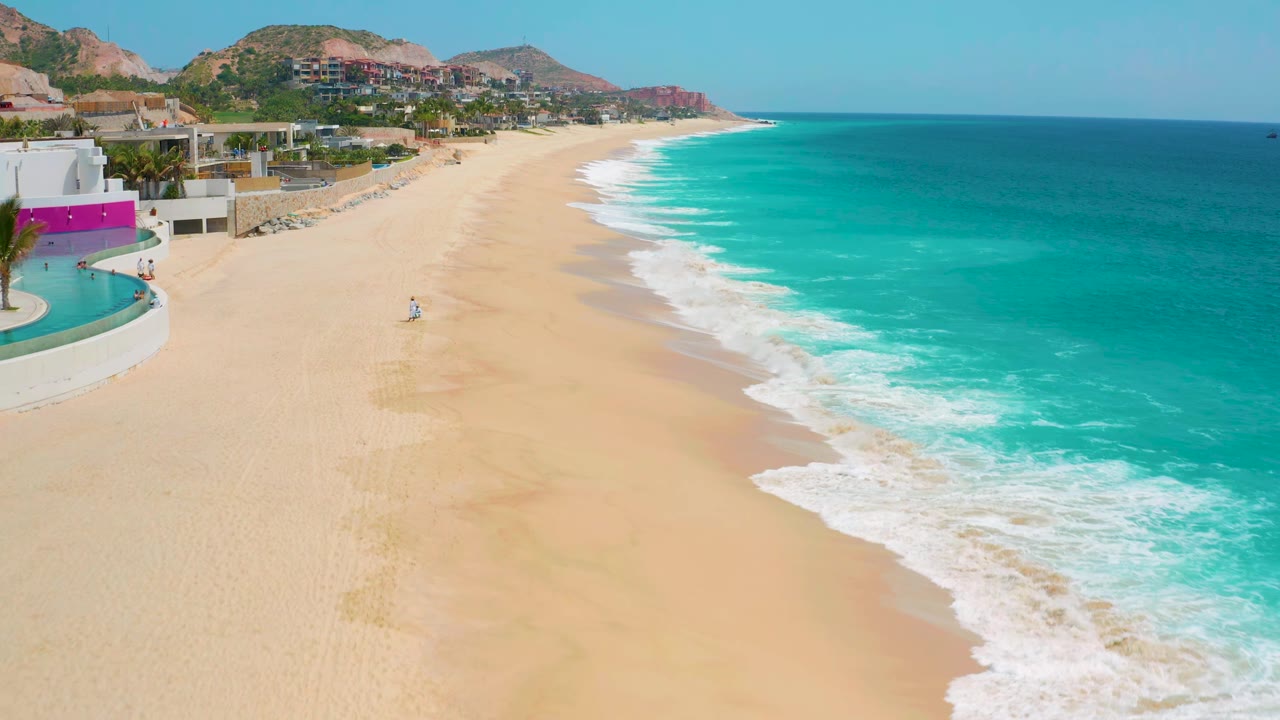 Wide aerial-style frame of Secrets Marquis Los Cabos beach, with a curved band of foam at the waterline, resort structures at left, hills beyond, and a small beachgoer near the center of the sand.