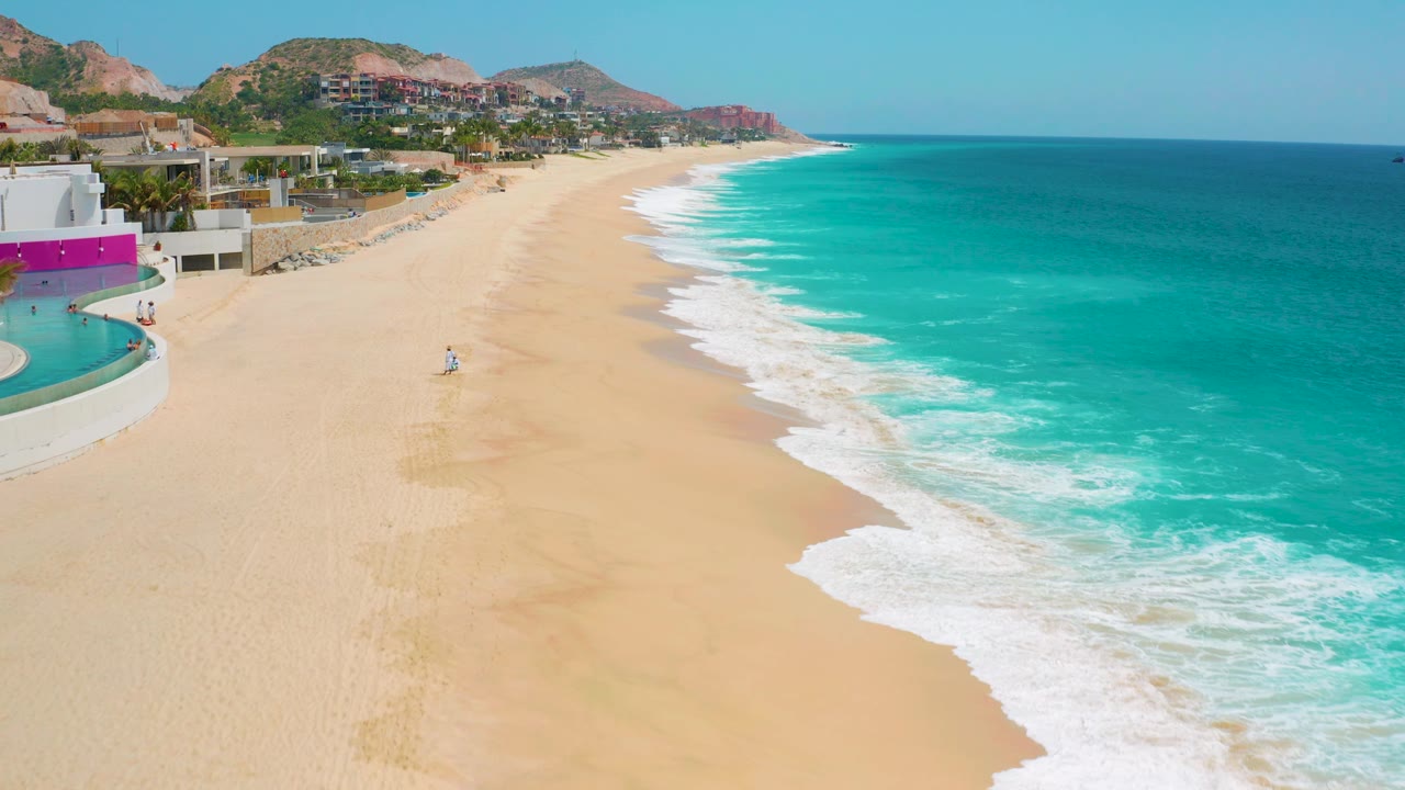 The shoreline at Secrets Marquis Los Cabos fills the frame with pale sand on the left, bright turquoise water on the right, low hills in the background, and one distant person on the beach.