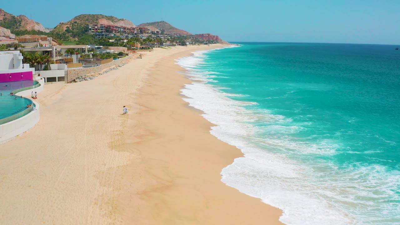 Sandy coast with white waves rolling in beside resort buildings and hills.