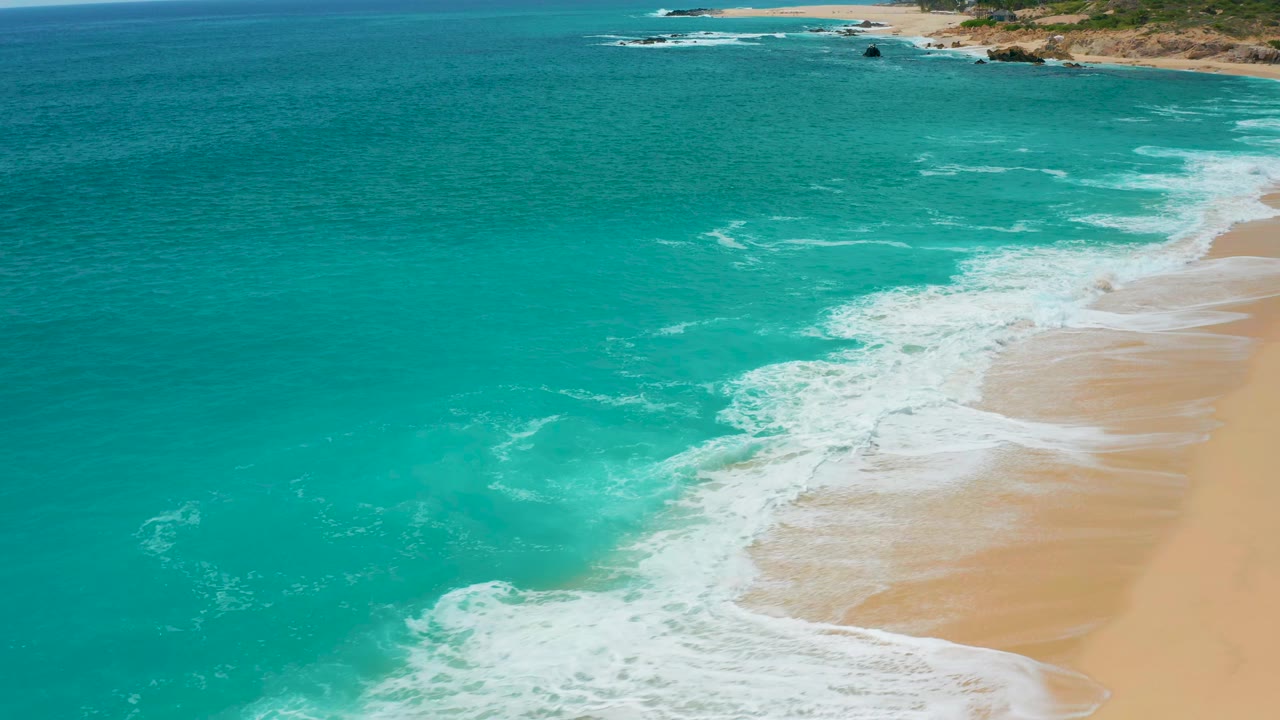 Aerial view at Secrets Marquis Los Cabos, Mexico, where a broad band of white foam reaches farther up the sandy shoreline with no visible people.