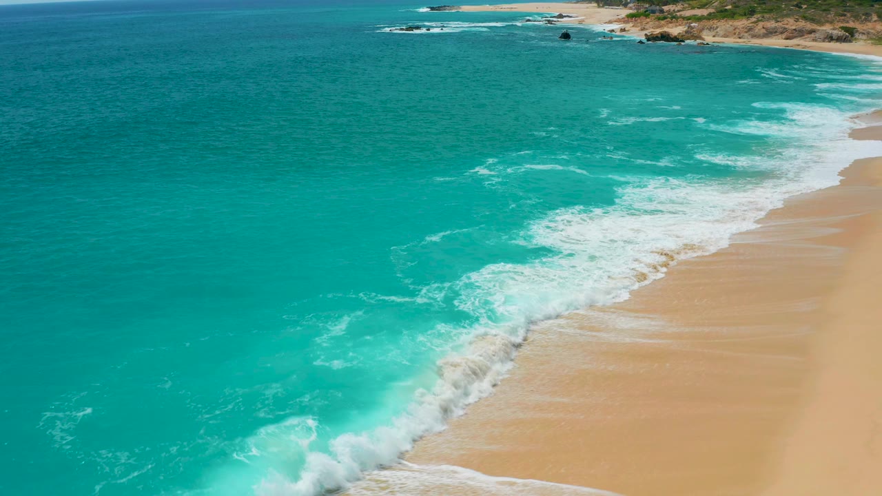 Elevated view over Secrets Marquis Los Cabos, Mexico, showing a thicker white break of surf along the right-side beach and no visible people.