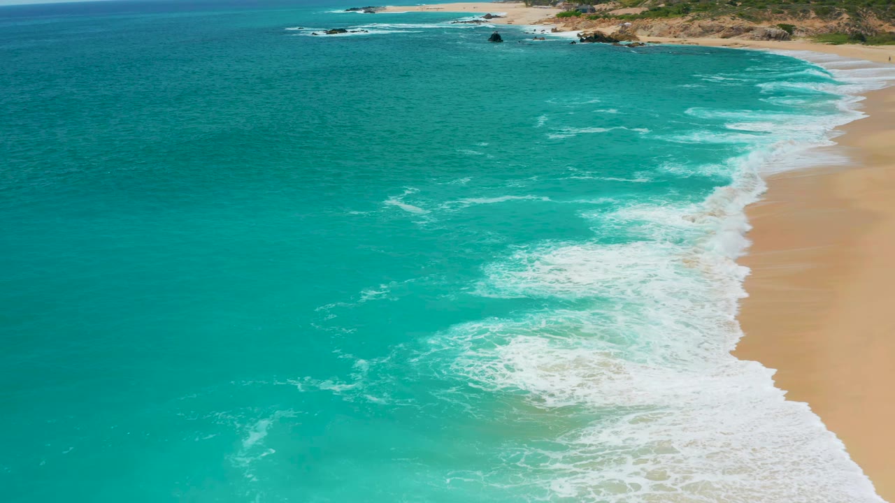 Aerial shoreline frame at Secrets Marquis Los Cabos, Mexico with a broad teal water field, fresh white foam curling toward the beach on the right, and no visible people.