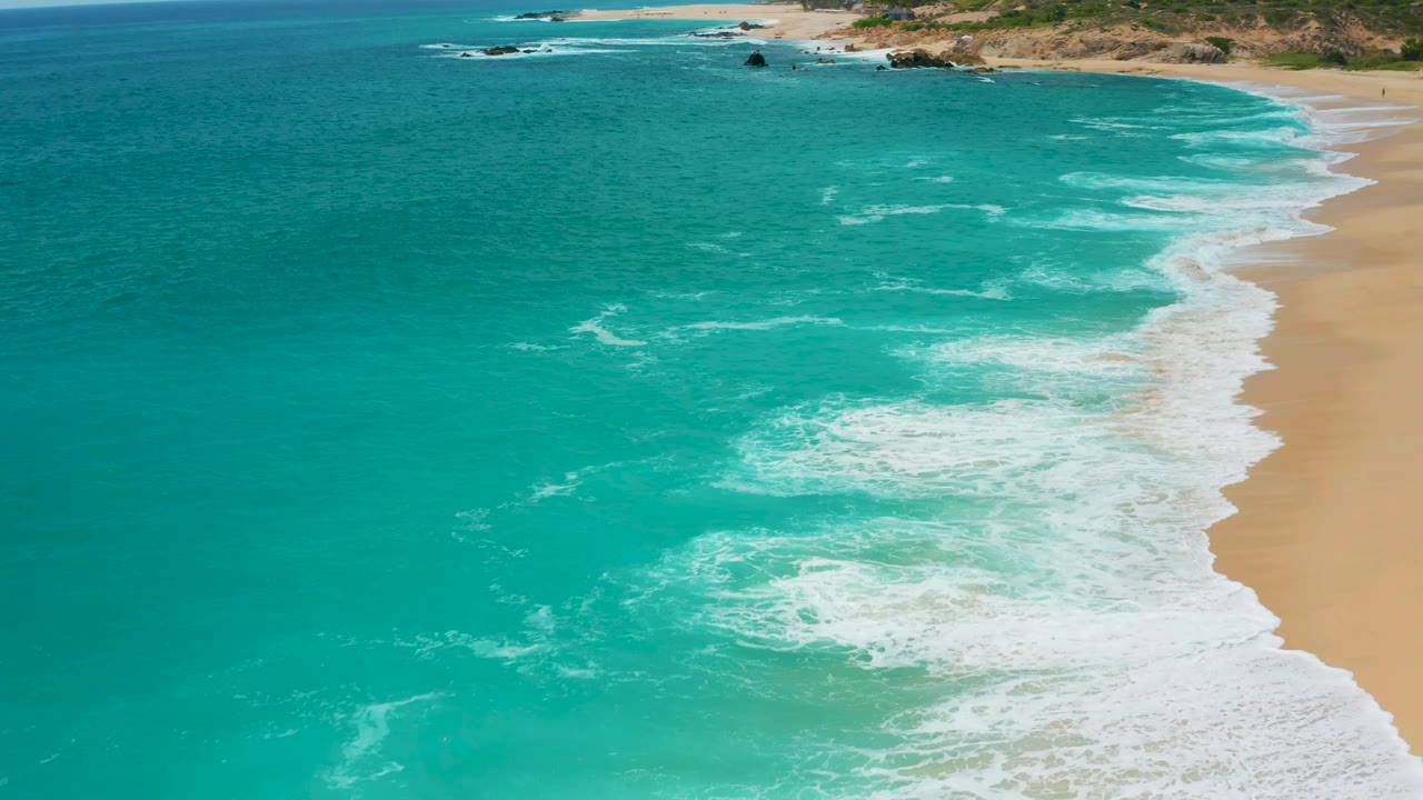 Elevated coastal view at Secrets Marquis Los Cabos, Mexico where broken foam spreads across turquoise water beside the beach, with rocky land across the upper frame and no visible people.