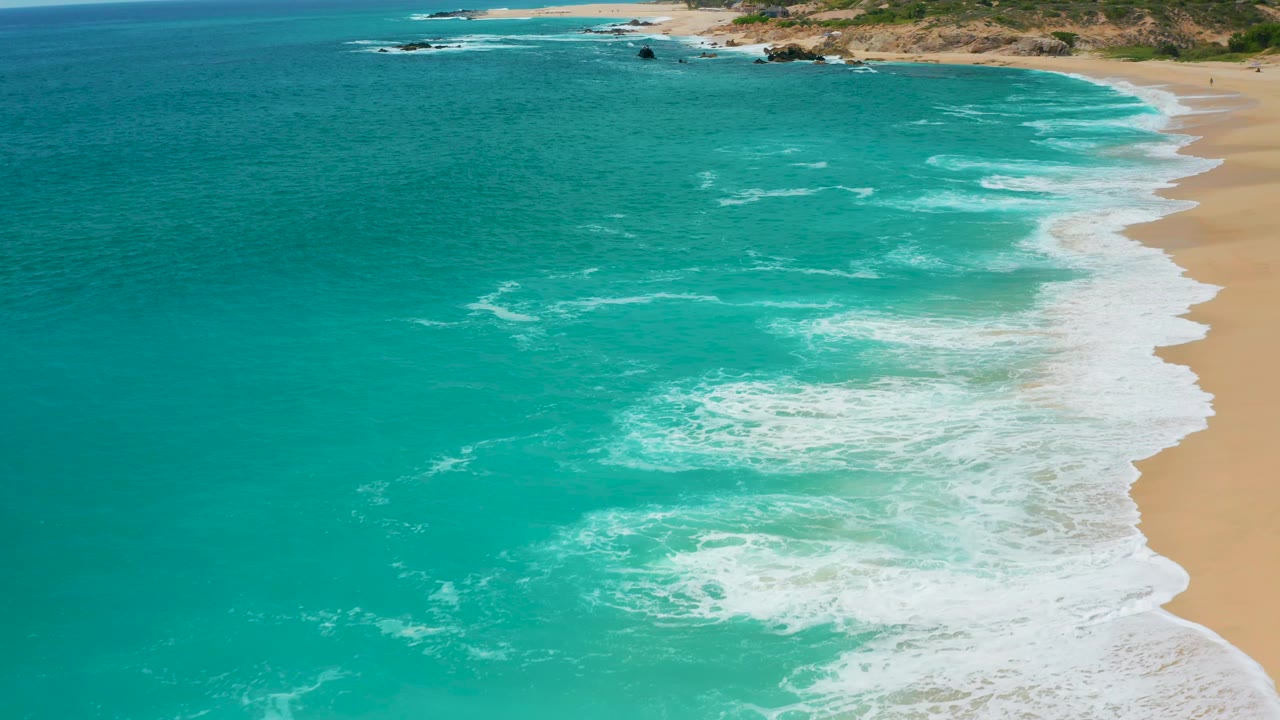 Secrets Marquis Los Cabos, Mexico shoreline seen from above, with white surf tracing a diagonal edge between teal ocean and tan beach, rocky coast in the distance, and no visible people.