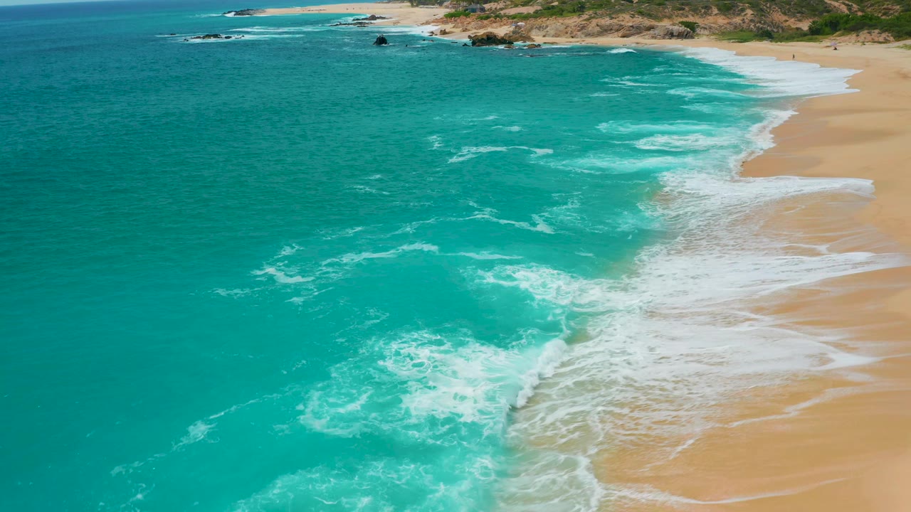 Aerial view of the Secrets Marquis Los Cabos, Mexico shoreline with blue-green water on the left, fresh surf on the right, and no visible people on the beach.