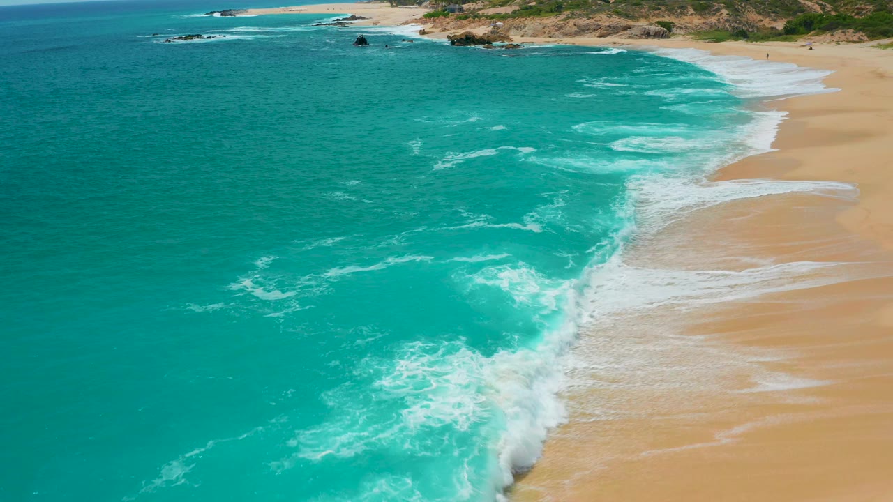 Aerial frame at Secrets Marquis Los Cabos, Mexico, showing a diagonal line of white surf washing onto empty tan sand, with rocky coast in the distance.