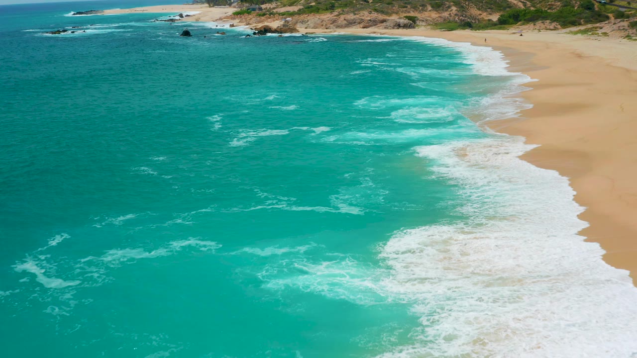 Aerial beach frame from Secrets Marquis Los Cabos, Mexico, with foamy surf spreading through shallow turquoise water beside open sand, no visible people.