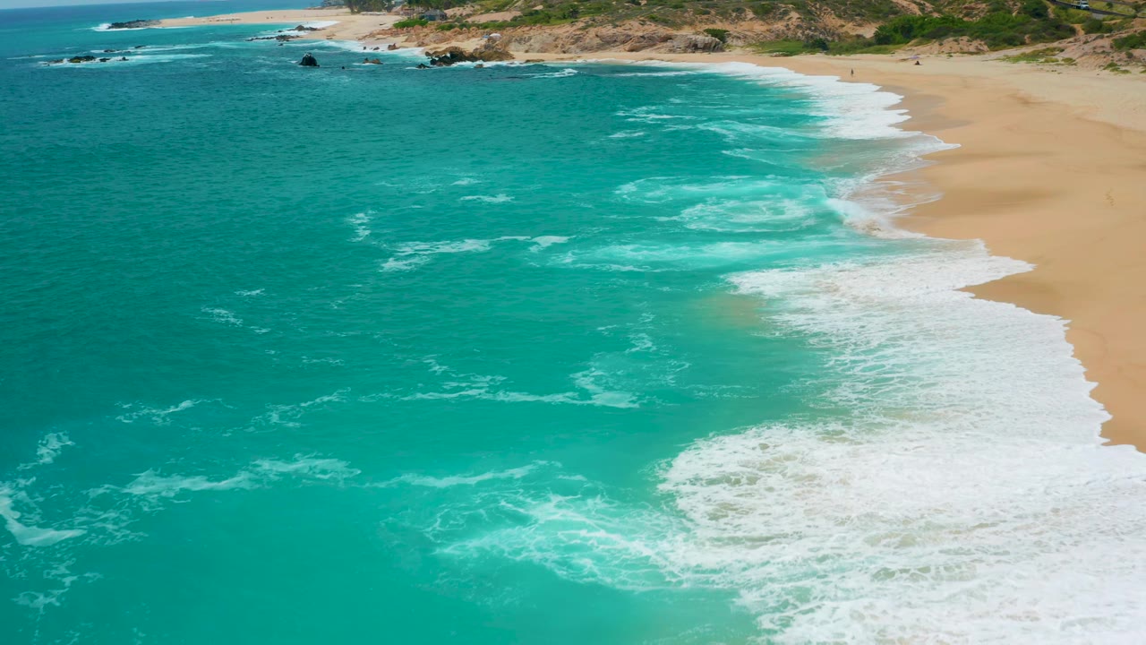 High aerial view at Secrets Marquis Los Cabos, Mexico, with a curved white surf band along the sandy shoreline and rocky coast in the distance, no visible people.