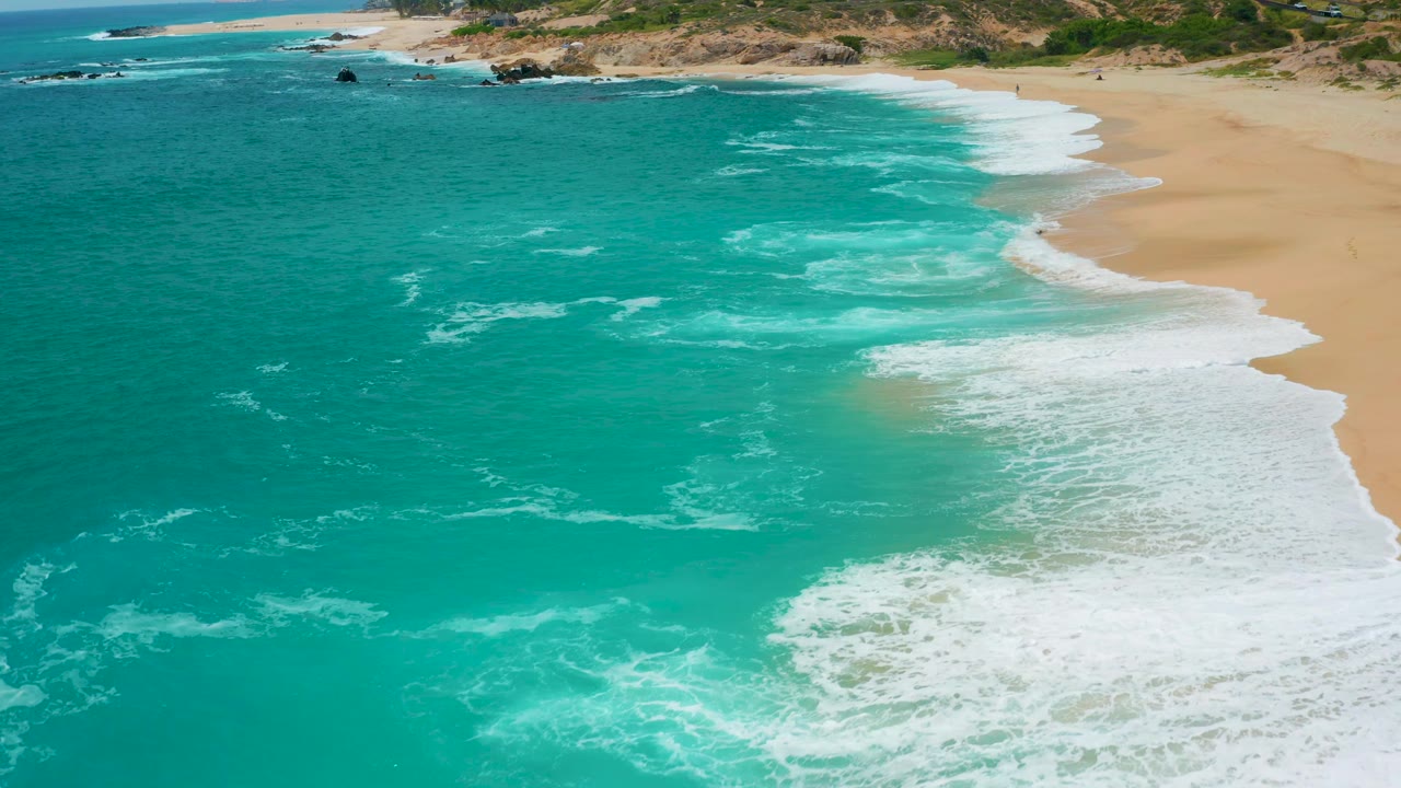 Aerial view over Secrets Marquis Los Cabos, Mexico, showing teal water in the foreground, white foam on the right, tan sand above it, and rocks near the far shoreline, no visible people.