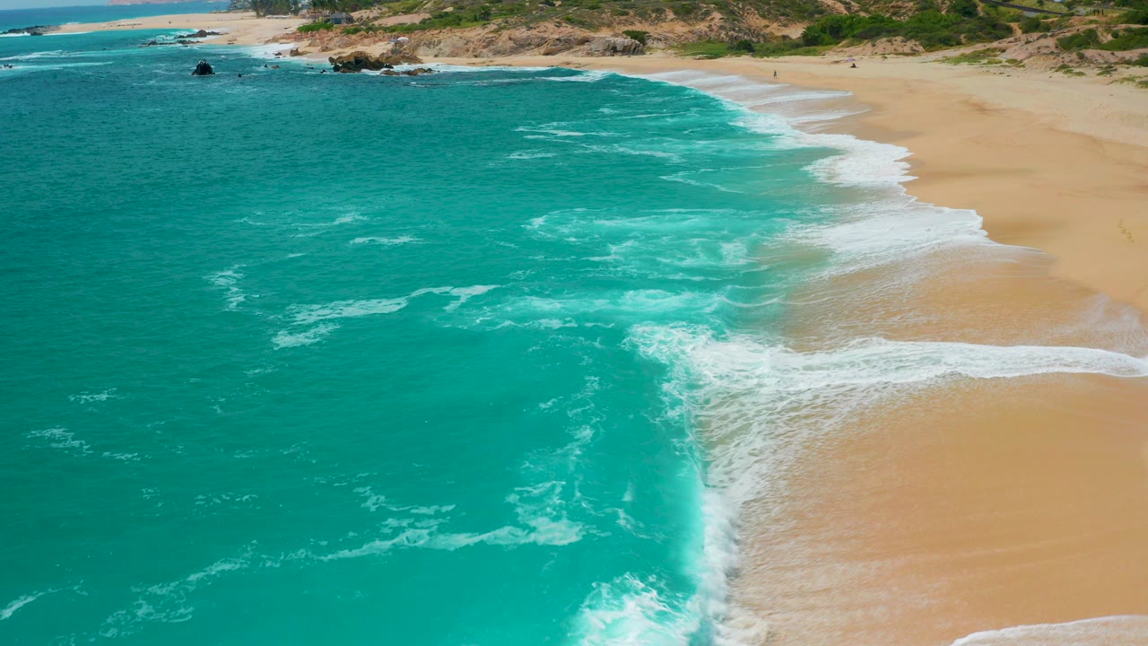 Aerial view at Secrets Marquis Los Cabos, Mexico, with turquoise ocean on the left, white surf curling onto tan sand, low dunes beyond the beach, and no visible people.
