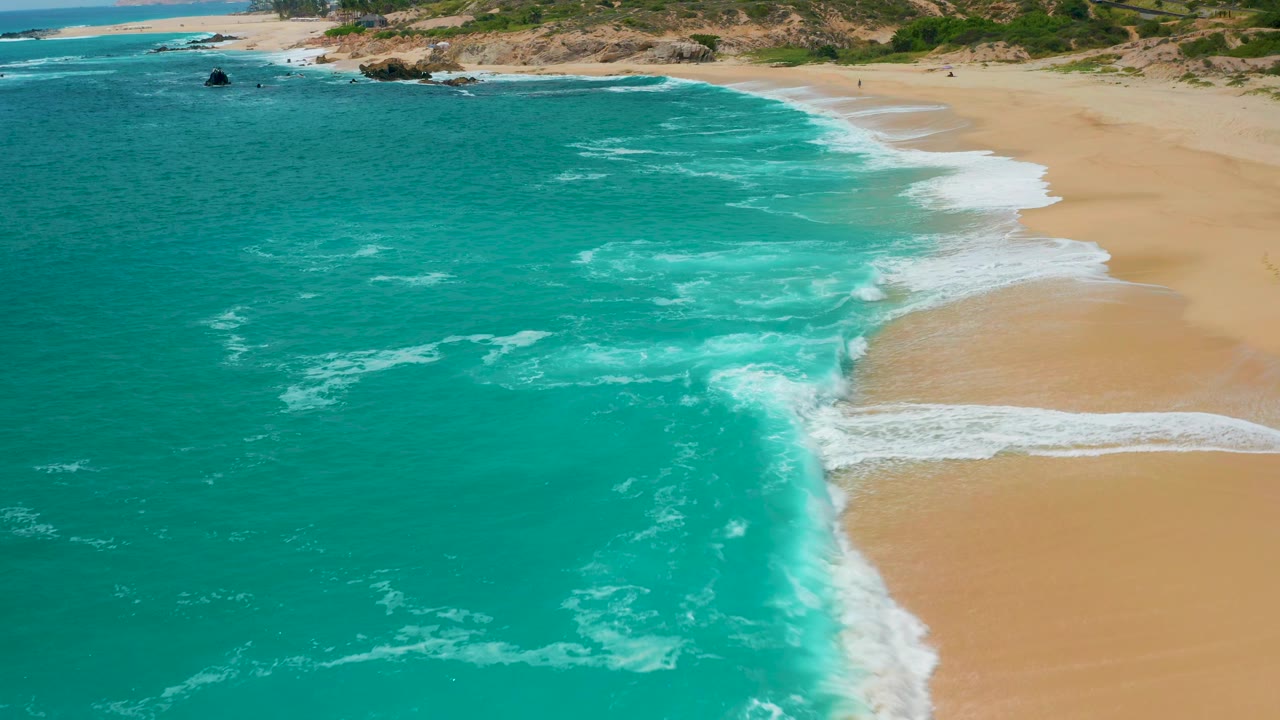 Aerial view at Secrets Marquis Los Cabos, Mexico, with a curved white wave edge meeting broad tan sand, turquoise water filling most of the frame, rocky coast in the distance, and no visible people.