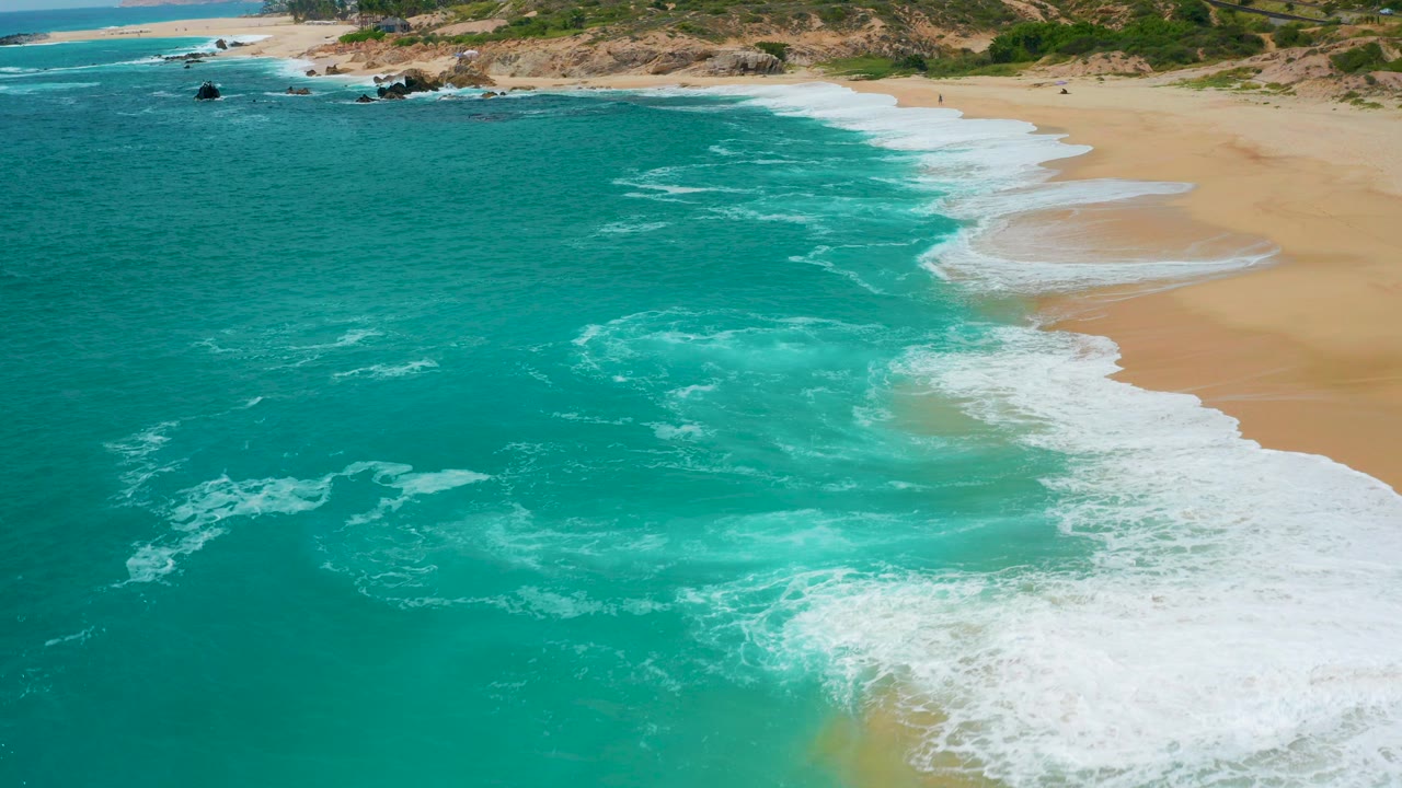 Aerial view at Secrets Marquis Los Cabos, Mexico, with turquoise ocean filling the left side, white surf reaching tan sand, dune scrub beyond the beach, and no visible people.