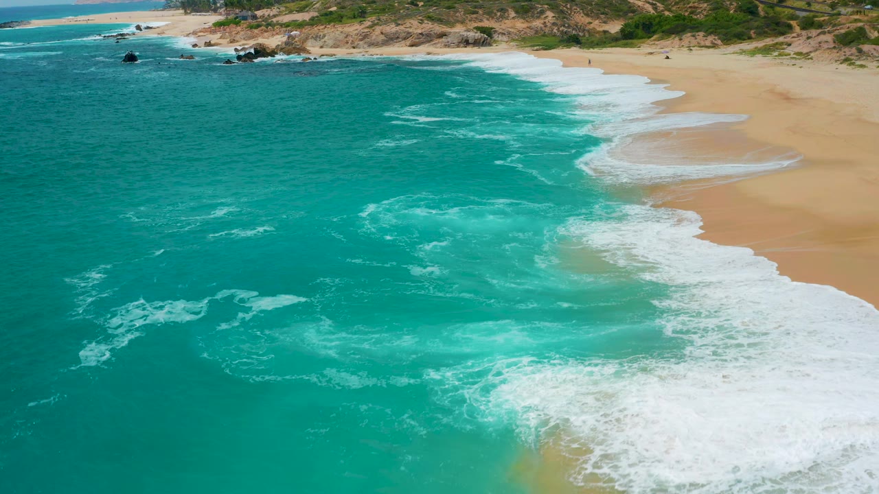 Aerial coastal frame from Secrets Marquis Los Cabos, Mexico, showing a broad patch of white foam in turquoise water, wet sand along the right edge, scrub-covered dunes above, and no visible people.