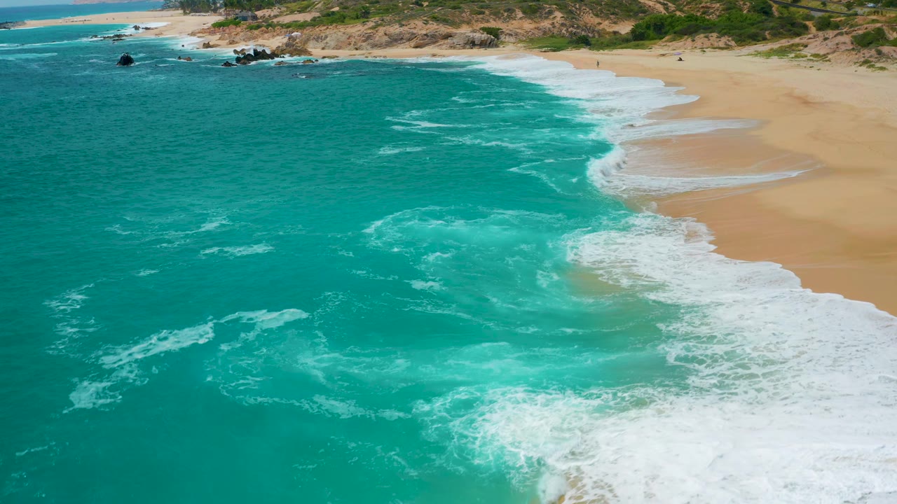 Secrets Marquis Los Cabos, Mexico shoreline seen from above, with deeper teal water at left, white breakers curling into the sand at right, scrubland above the beach, and no visible people.