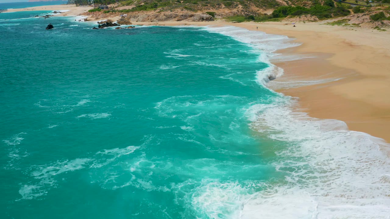 Aerial view over Secrets Marquis Los Cabos, Mexico, where turquoise water covers most of the frame, white foam gathers near the lower shoreline, tan beach runs along the right side, and no visible people.