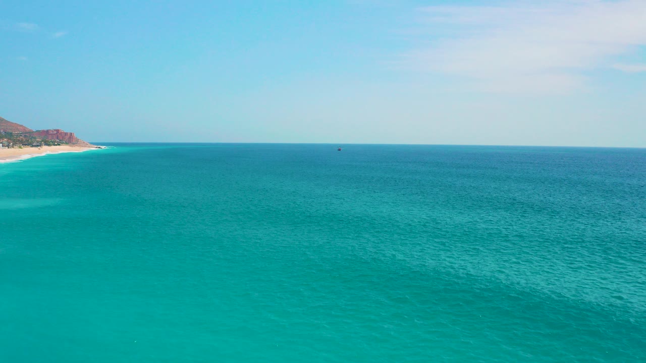 Wide turquoise ocean near Secrets Marquis Los Cabos, Mexico, with a pale beach and low brown headland on the far left under bright daylight, no visible people.